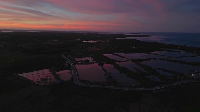 Elevated wide aerial capturing rice fields, rural road, and glowing sunset sky over Tondol, Anda countryside.