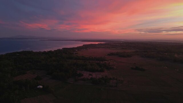 Drone pulls back from a quiet road revealing wide rice fields glowing under a dramatic sunset sky in Tondol.