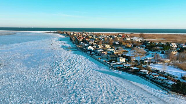 A stunning drone shot of the frozen Baltic Sea coastline in Rewa, Poland, featuring snowcovered houses and icy waters under a clear blue sky.