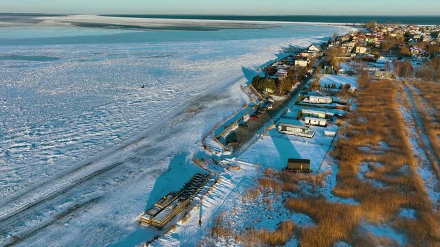 High angle aerial shot of a snow covered beach and frozen sea ice next to a small coastal village in Rewa, Poland during winter sunset.