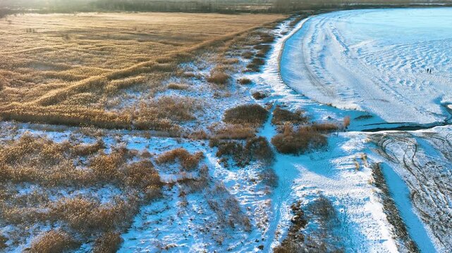 Stunning aerial shot of a frozen coastline in Rewa, Poland, featuring golden reeds and snowcovered ground during a cold winter sunset.