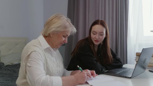 Senior woman in white blouse writes notes while teen girl in black hoodie uses laptop at white desk in home interior, symbolizing family mentoring and education.
