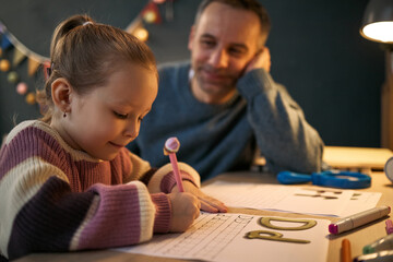 Fototapeta premium Caucasian child girl practicing writing letters on worksheet while middle aged Caucasian man watching attentively, both sitting at desk with educational materials in cozy indoor setting