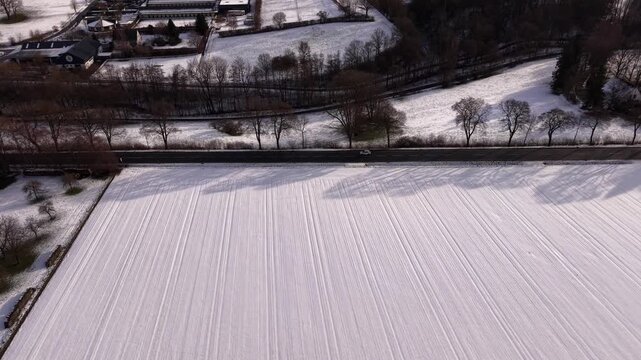 Driving car on snow-covered roads winding through town, with fields and buildings dusted in white. Warsteiner Brewery complex is visible in background. Aerial tracking shot. Germany in winter.