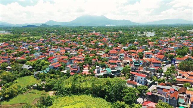 Drone shot over Duong Lam ancient village in Vietnam, showing traditional red-tiled roofs, lush green vegetation, and the majestic Ba Vi mountain range in the background under a bright sky