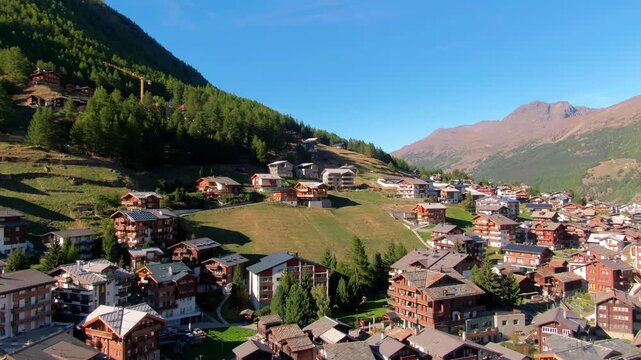Stunning aerial view of Saas-Fee, Switzerland &ndash; a vibrant alpine village with charming chalets, lush green hillsides, dense forests, and majestic snowy peaks under a clear blue sky