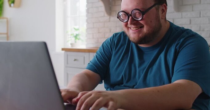 Joyful overweight funny man typing working on laptop at home. Wearing round glasses, he reacts with surprised smile to pc in kitchen. Humorous energy and positive online fun vibes.