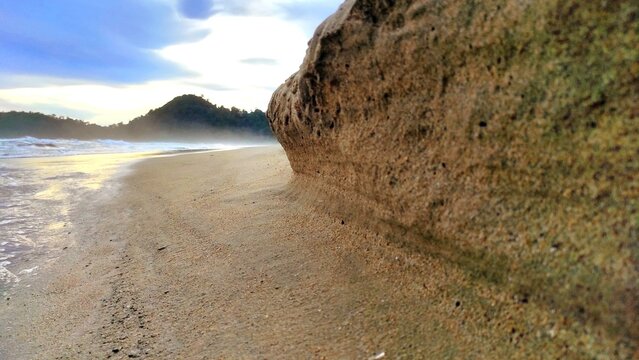 Low angle view of a natural rock formation on a sandy beach with ocean waves and distant hills under a soft sky, creating a calm and coastal atmosphere