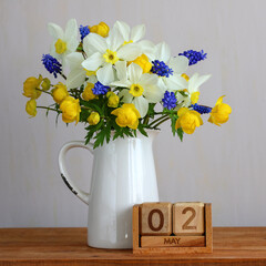 Naklejka premium spring bouquet of daffodils, muscari, and trollius in a white pitcher standing on a wooden surface next to a vintage calendar displaying May 2nd, symbolizing seasonal renewal