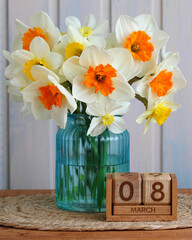 Daffodils in a teal ribbed vase on a woven mat with a wooden perpetual calendar showing March 8, representing International Women's Day and arrival of spring