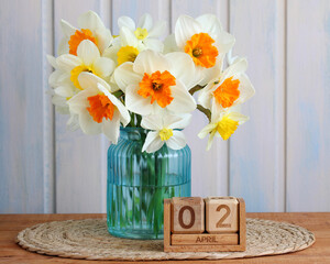 daffodil flowers displaying white petals and orange crowns blooming in a turquoise glass vase next to a wooden block calendar showing April 2nd, representing spring and new beginnings
