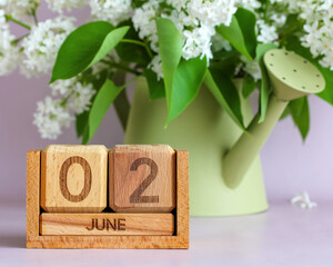 Wooden perpetually changing calendar displaying June 2, nestled near a light green watering can holding white lilac flowers, representing spring and early summer blooms