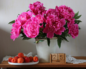 Peonies blooming in a white bucket, apricots on a white plate, and a wooden perpetual calendar showing June 25, creating a serene summer still life