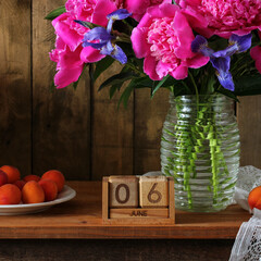 Vase of vibrant pink peonies and blue irises on a rustic wooden shelf with a June 6 block calendar and fresh peaches in a still life setting, symbolizing summer and special dates