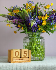 Wildflowers bouquet with purple lupine and yellow buttercups in a glass vase, standing next to a block calendar displaying June 5, symbolizing nature, summer, and the World Environment Day