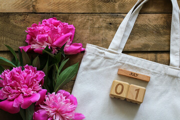 Vibrant pink peonies are blooming in summer, arranged next to a blank canvas tote bag and a wooden block calendar displaying the date July 01 on a rustic wooden background
