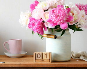 Peonies bouquet in a rustic milk pitcher on a wooden table with a perpetual calendar set to July 1, pink mug and lace creating a cozy, romantic summer morning still life