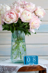 Peonies in a glass vase beside a wooden block calendar showing March 8, soft pastel still life celebrating International Women's Day, spring blooms and feminine grace