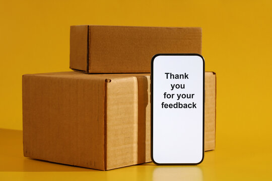 Smartphone displaying a thank you for feedback message next to stacked cardboard delivery boxes on a yellow background, symbolizing customer satisfaction for e commerce and logistics services