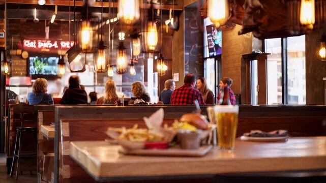 A cozy restaurant scene with a main table in foreground featuring a burger, fries, and a pint of beer, while warm pendant lights glow overhead and blurred diners sit at booths in the background.