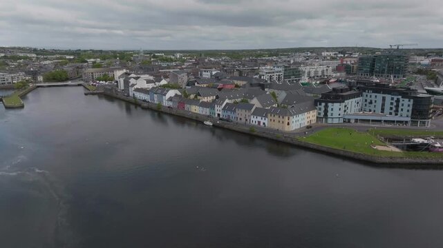 A cinematic drone shot capturing the colorful houses of Long Walk and the calm River Corrib in Galway, Ireland under a cloudy sky.