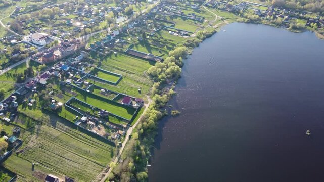 Drone view of a charming village descending to a calm lake, showcasing a Orthodox temple amid vibrant spring greenery and peaceful homes. Kaluga region, Russia