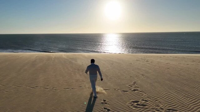 Man in White Walking on Sand Dunes Towards the Ocean at Sunset