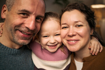 Fototapeta premium Portrait of Caucasian middle aged man, Caucasian woman, and Caucasian child girl smiling closely together, child girl hugging adults, all looking into camera