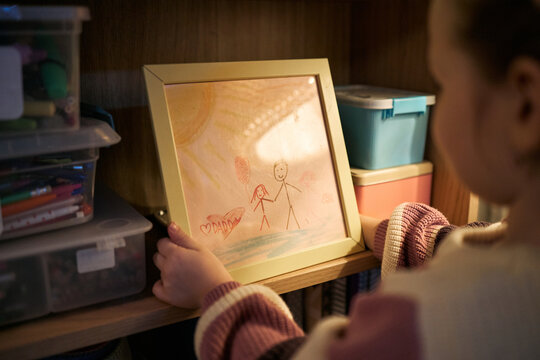 Caucasian child holding framed drawing depicting family under sun, standing near shelf with art supplies, showing back of head and hands while looking at picture