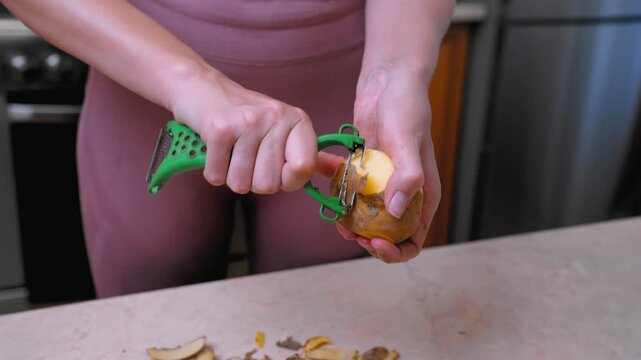 Ingredient preparation scene. Hand peeling potato carefully. Closeup of hands preparing ingredients leisurely. Someone skillfully peeling potato with peeler on kitchen countertop