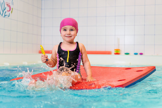 Happy little girl splashing water sitting on swimming board in pool