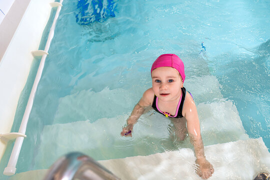 baby girl in a pink swim cap standing in the pool water.