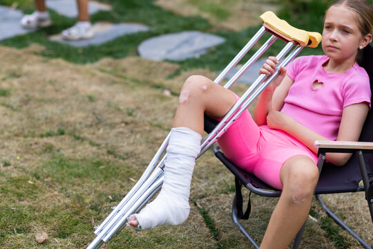 Little caucasian girl walking outdoor with plaster bandage on foot. Bone fracture foot and leg on child patient with splint cast and crutches during surgery rehabilitation and orthopedic recovery