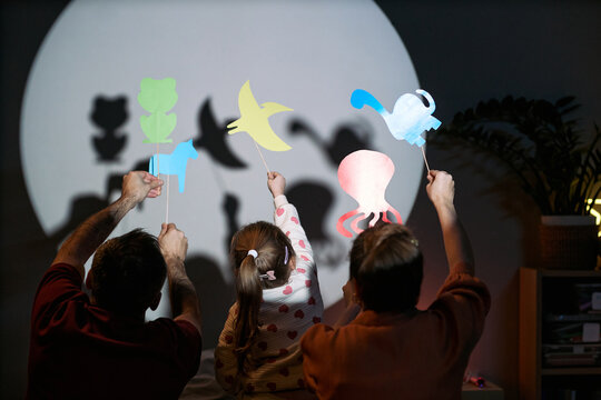 Caucasian man, Caucasian woman, and Caucasian child holding colorful paper shadow puppets in front of illuminated wall, creating animal shapes with hands raised together