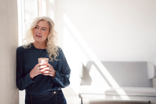 Mature woman enjoying coffee break by window in sunny modern loft