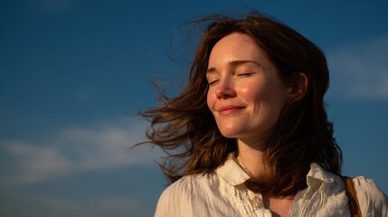 Fototapeta premium A woman with closed eyes enjoys a gentle breeze, basking in warm sunlight against a clear blue sky.