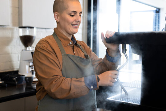 Female barista wearing grey apron using steam wand at cafe counter with grinder, cups, pitcher
