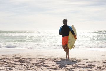 Obraz premium Full length rear view of senior woman running with surfboard towards sea at sunny beach