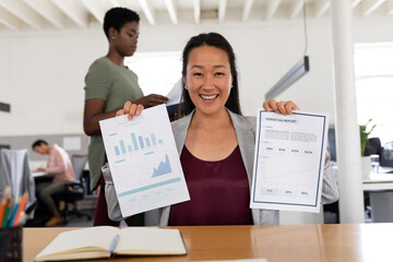Portrait of smiling asian creative businesswoman showing charts while sitting at desk in office