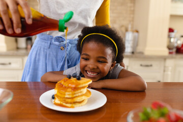 Mother pours syrup on daughter's pancake, showcasing family love.