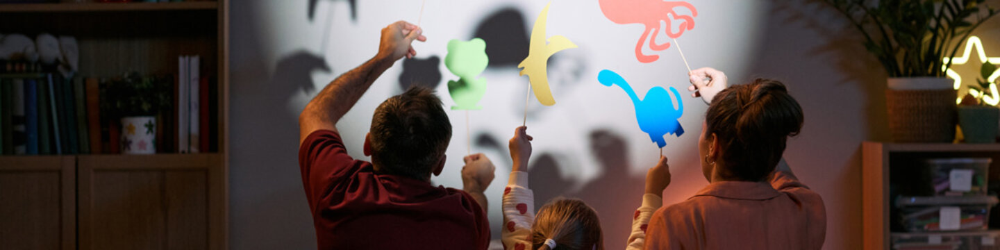 Caucasian man, Caucasian woman, and child creating animal shadow puppets on wall, raising colorful paper cutouts together, backs facing camera, engaging in playful family activity