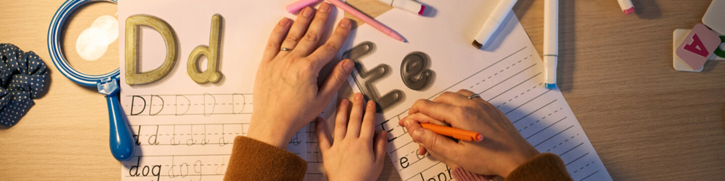 Caucasian adult guiding Caucasian child in practicing handwriting on worksheet, both hands holding pencil together, foam alphabet letters and markers scattered on wooden desk