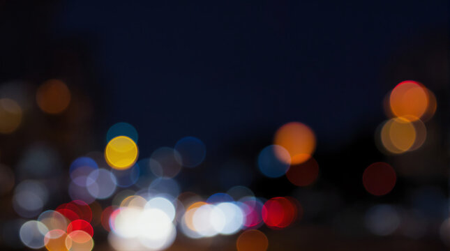 Blurred city lights create a colorful bokeh effect against a dark nighttime background, highlighting vibrant reds, blues, yellows, and whites in soft, circular shapes