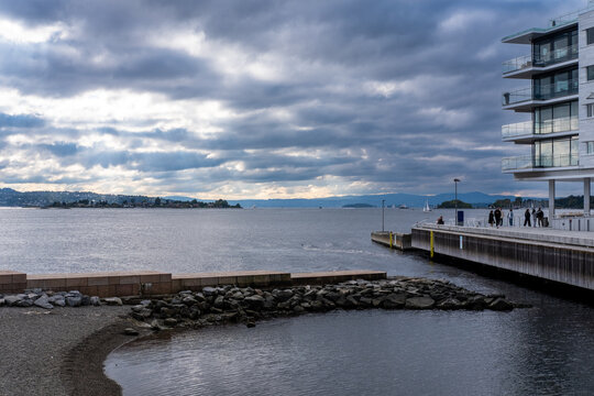 Overcast waterfront shoreline with rocks by fjord and architecture in urban Oslo Norway featuring clouds calm water and Nordic city landscape