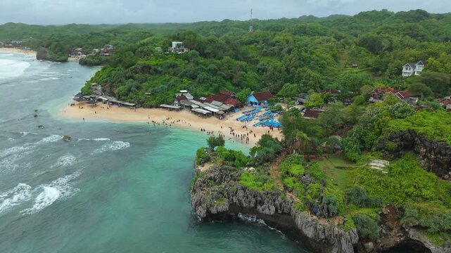 Aerial view of Ngandong Beach where turquoise waters meet the sandy shore, with lush green hills rising in the background, Gunungkidul Regency, Indonesia.
