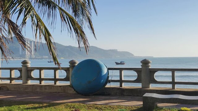 Traditional Vietnamese Coracle rests by concrete railing at sunny coastal promenade. Blue fishing boat and palm leaves create authentic atmosphere for Vietnamese Coracle display.