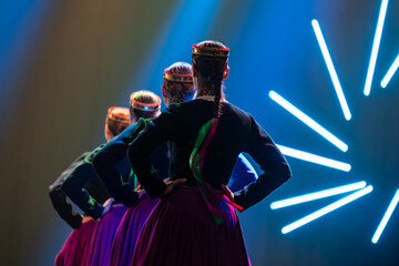 A line of dancers in traditional folk attire perform on stage, seen from behind, with braided hair and vibrant costumes under dramatic blue lighting