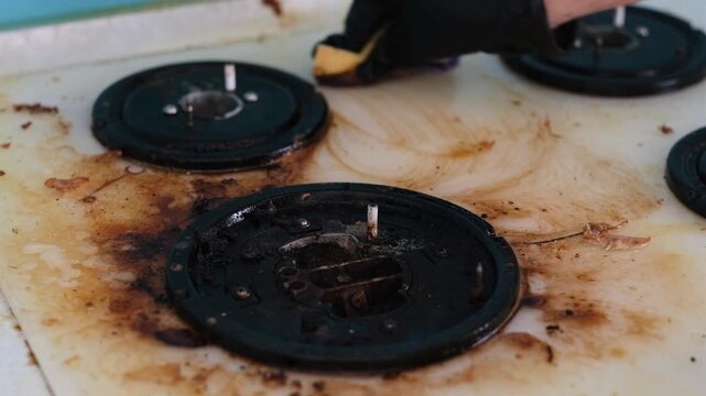 A woman's hand in a rubber glove washes a dirty gas stove.