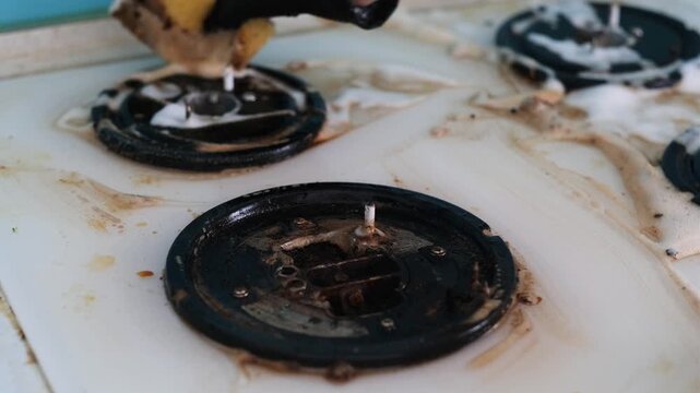 A woman's hand in a rubber glove splashes foam and washes a dirty gas stove.

