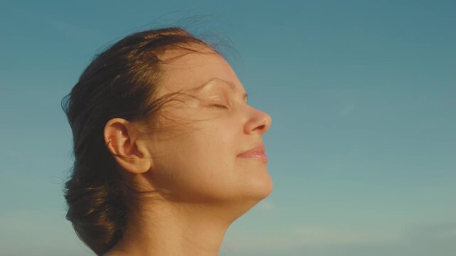 Woman enjoying golden hour sunlight on her face, feeling inner peace and freedom, relaxing outdoors with closed eyes, embracing mindfulness and wellness against a clear blue sky
